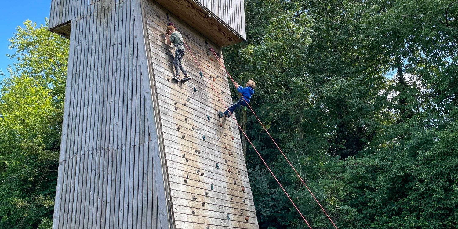 Students cheer as their peers scale a large wooden climbing wall.