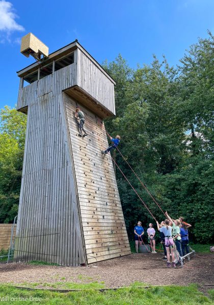 Students cheer as their peers scale a large wooden climbing wall.