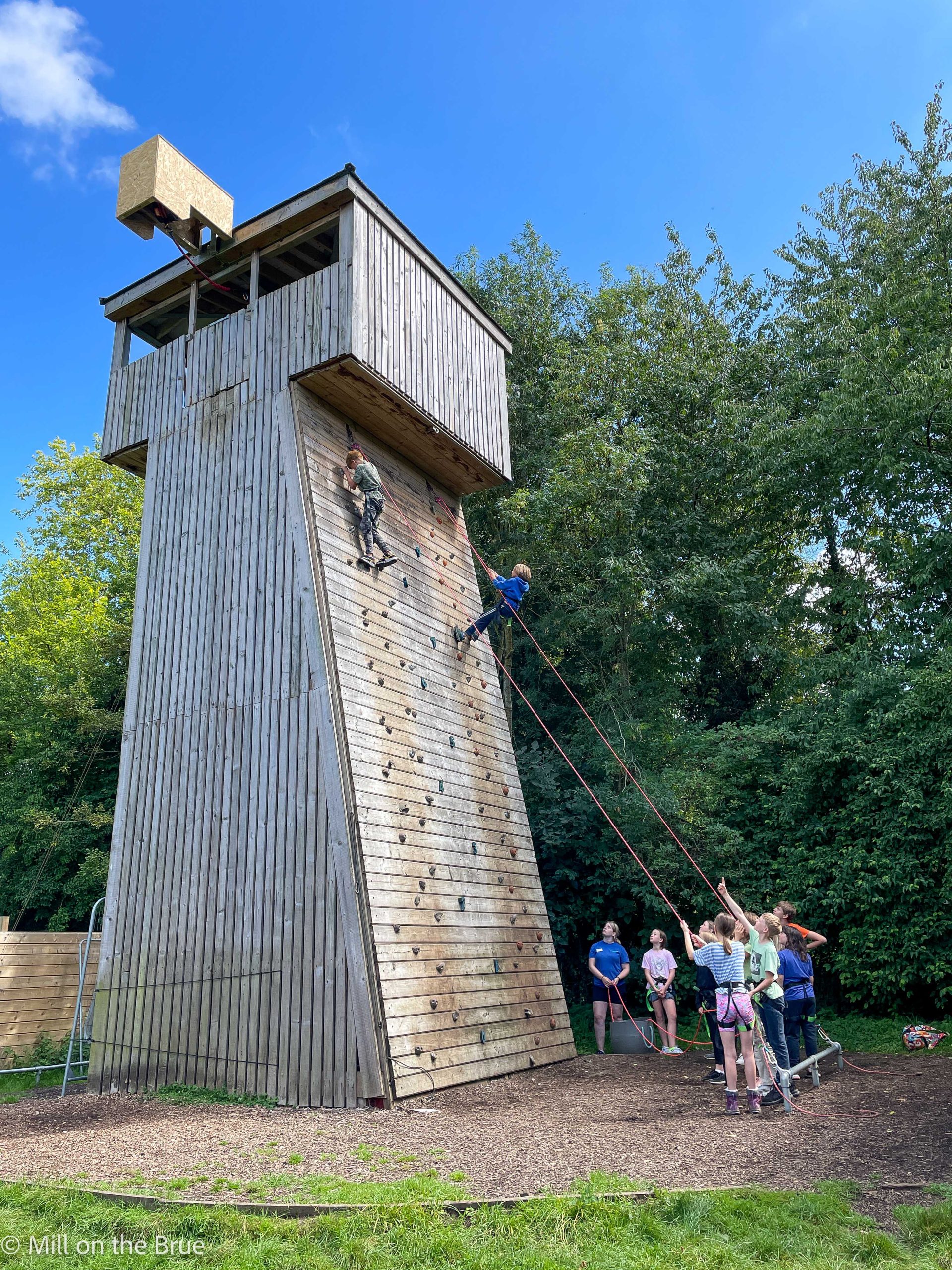 Students cheer as their peers scale a large wooden climbing wall.