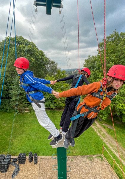 Students hold hands whilst suspended high in the air on ropes.